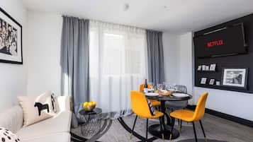 Een moderne woonkamer met een eethoek, een bank, een salontafel met een fruitschaal en een aan de muur bevestigde televisie waarop 'NETFLIX' wordt getoond.