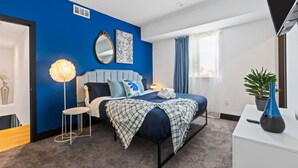 Ein modernes Hotelzimmer mit blauer Akzentwand, einem großen Bett mit vielen Kissen, einem Nachttisch mit Lampe und einer Topfpflanze.
