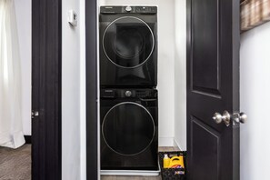 Una lavadora y secadora de color negro fijadas a la pared, con un panel de control, un termostato pequeño blanco y una botella de detergente amarilla en el suelo.