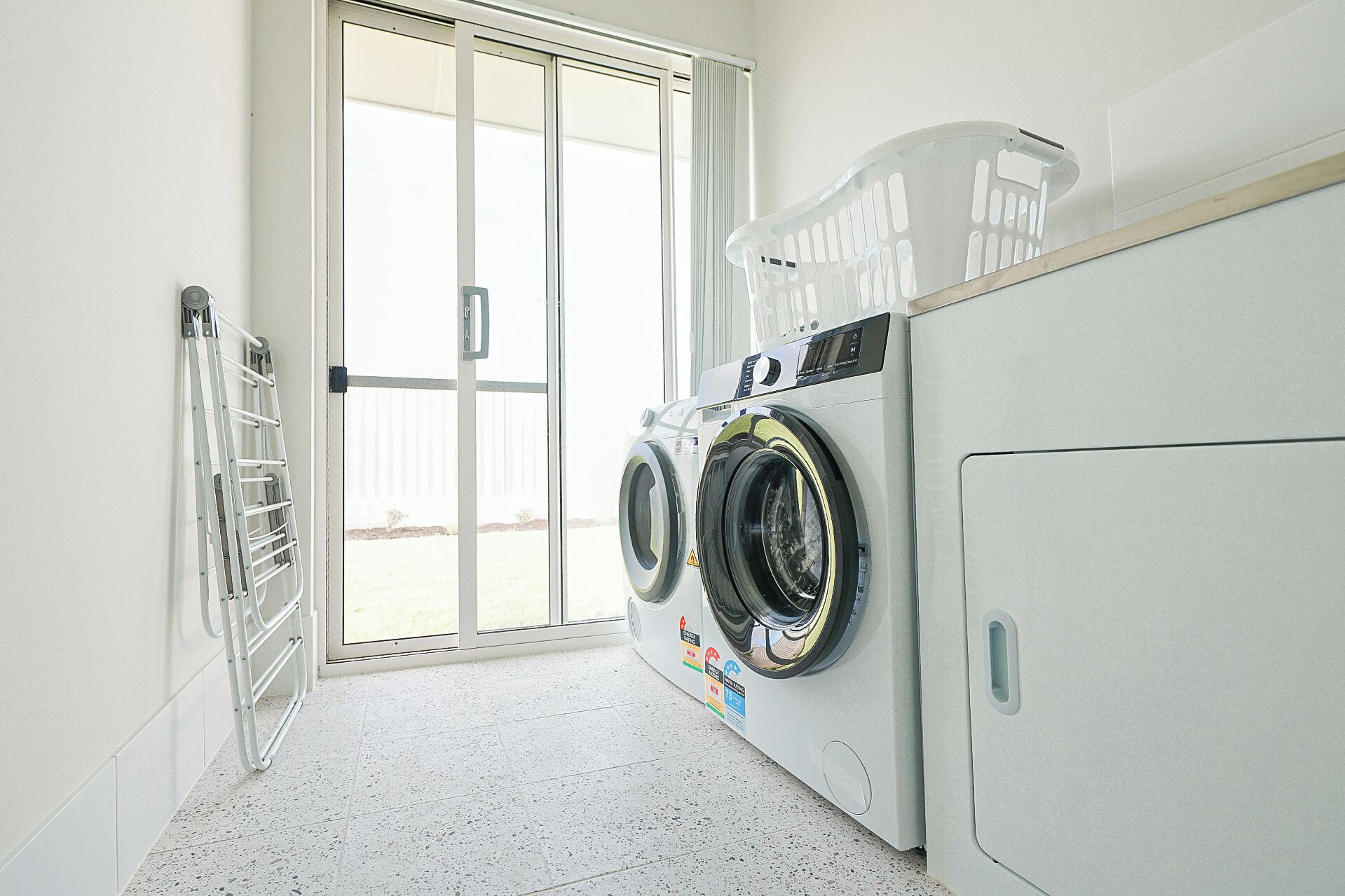 A laundry room with two front-loading washing machines, a drying rack, and a sliding glass door leading to an outdoor area.
