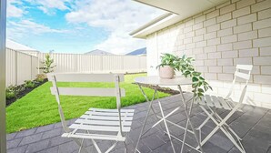 Un patio con una mesa y sillas blancas, una planta en maceta y vistas a un jardín con césped y un muro de ladrillo.