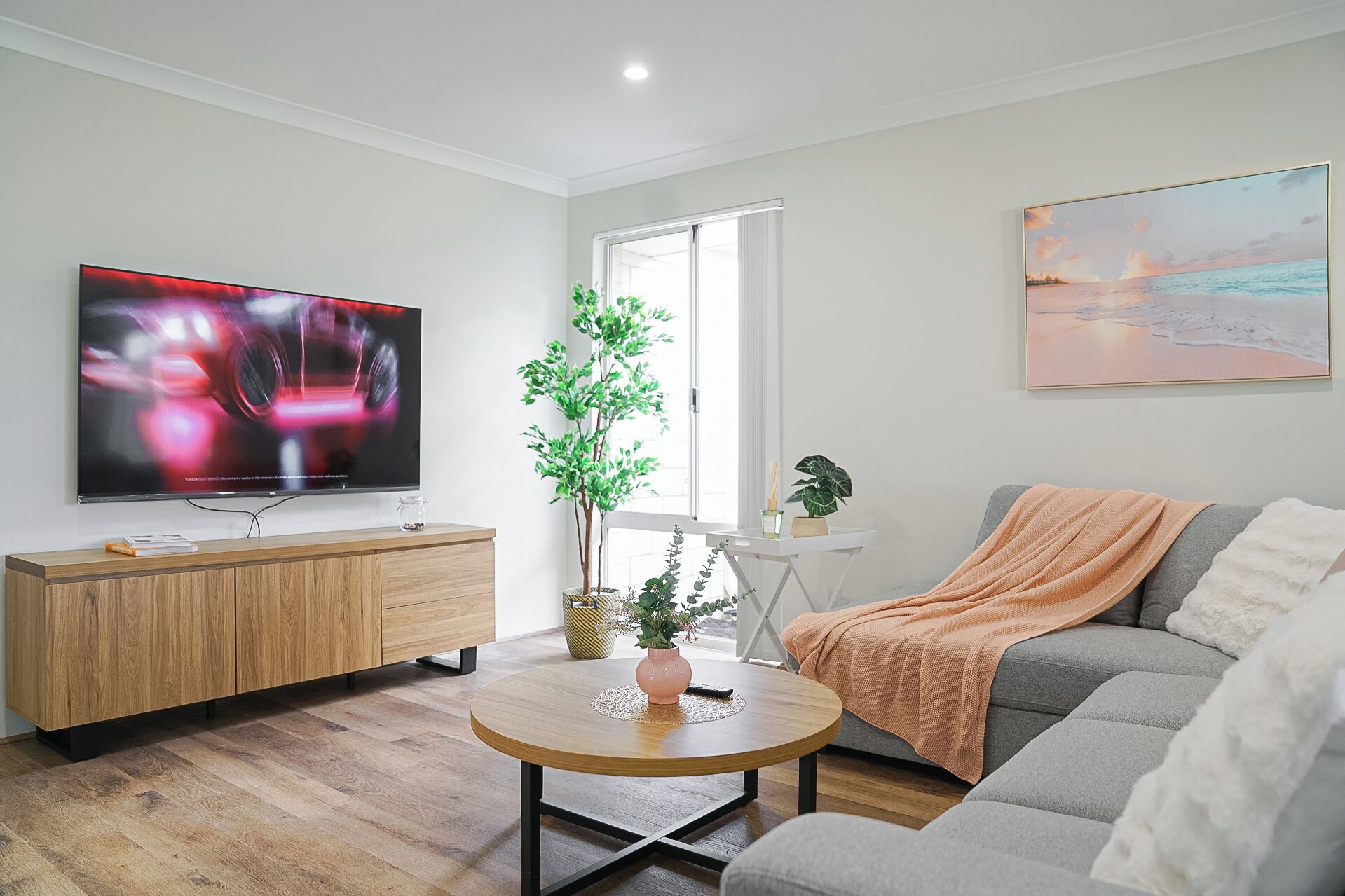 Una sala de estar moderna con un televisor de pantalla plana, un sofá gris, una mesa de centro redonda de madera y una mesita auxiliar blanca con una planta.