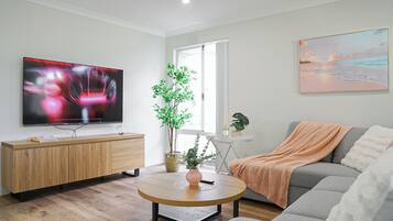 Una sala de estar moderna con un televisor de pantalla plana, un sofá gris, una mesa de centro redonda de madera y una mesita auxiliar blanca con una planta.