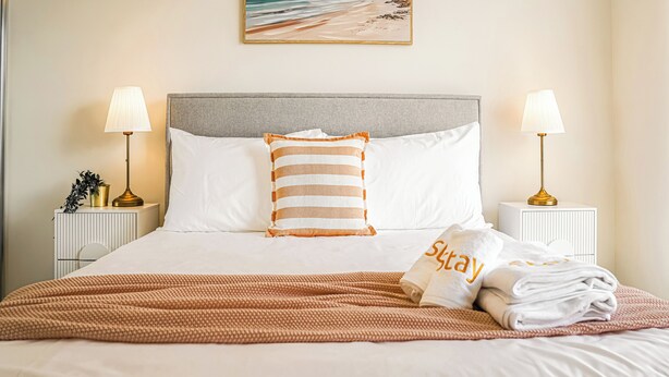 Una cama bien hecha con un almohadón a rayas, dos lámparas de noche y una toalla doblada con la palabra 'Stay'.
