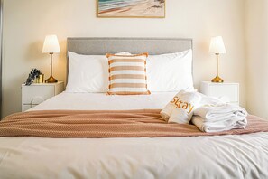 Een net opgemaakt bed met een gestreept kussen, twee bedlampen en een opgevouwen handdoek met de tekst 'Stay'.