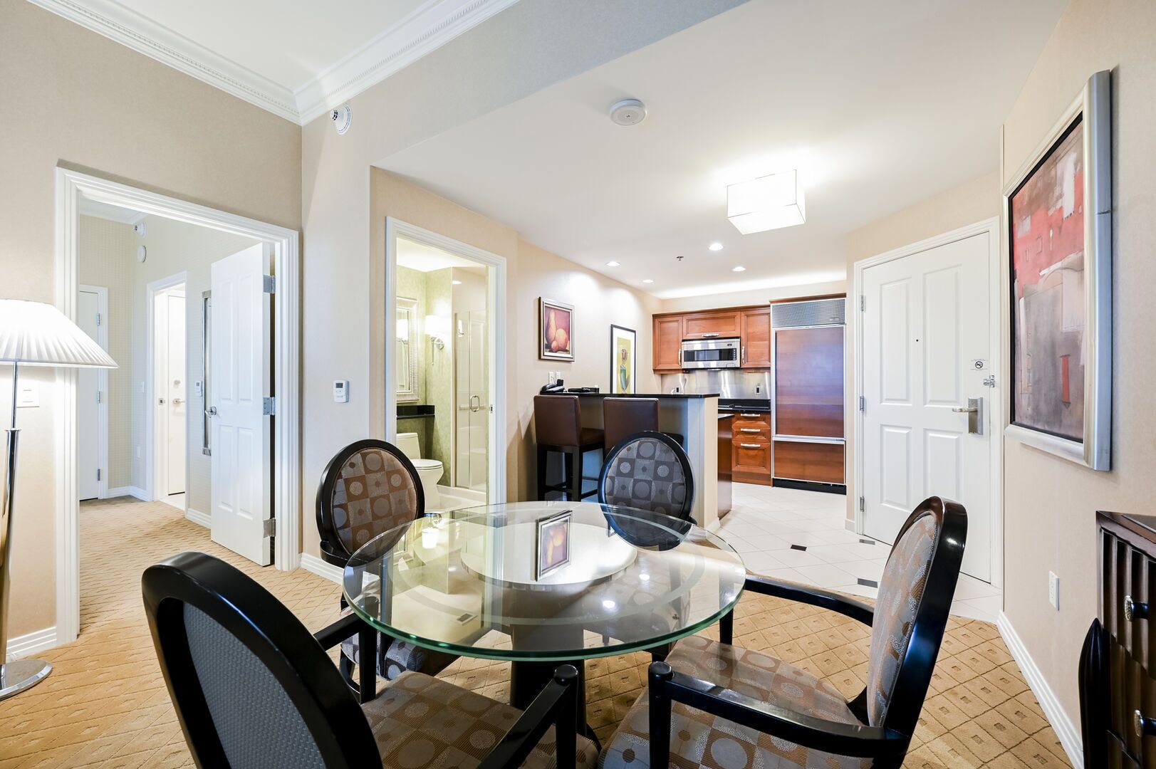 A dining area with a glass table and chairs, a kitchen in the background, and a hallway leading to a bathroom.