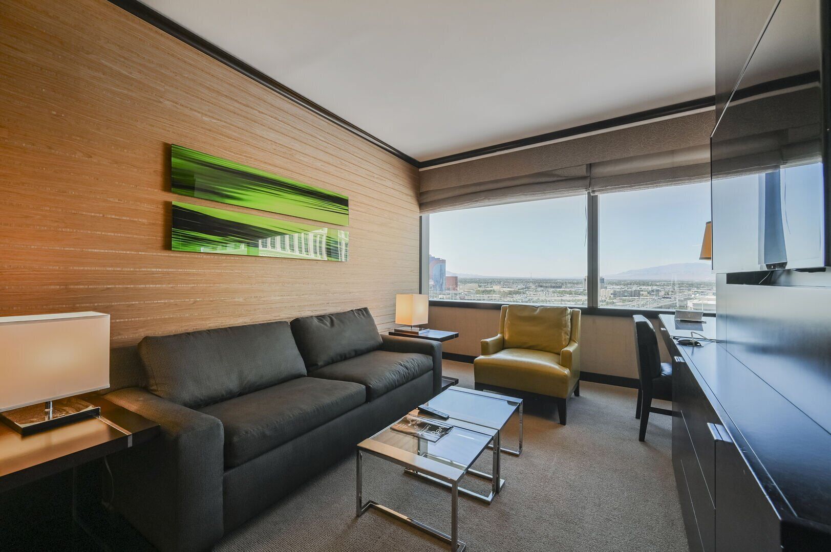 A modern hotel room with a sofa, armchair, coffee table, and a desk with a lamp.