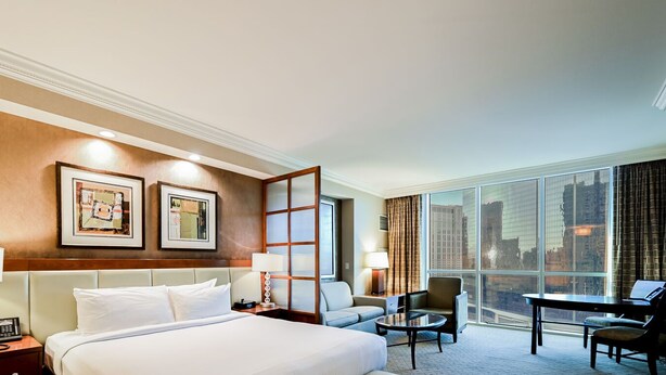 A hotel room with a large bed, two bedside tables with lamps, a desk with a chair, a sofa, and a large window with a city view. - SVS: 2BD Mountain View Suite | Private Balcony | Full Kitchen | MGM (Las Vegas)