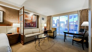 A hotel room with a sofa, armchair, coffee table, and a desk with a chair. There is a large window with a city view. - SVS: Penthouse 2BD | SPHERE Strip Views | Balcony | Signature at MGM (Las Vegas)