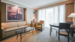Een hotelkamer met een bank, fauteuil, eettafel en een groot raam met gordijnen.