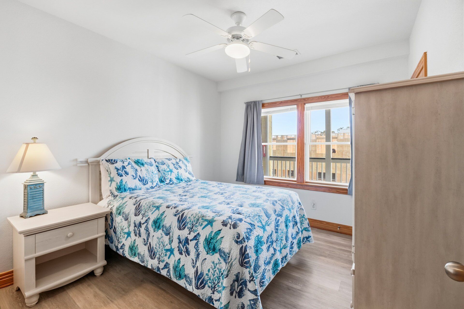 Un dormitorio con cama, mesita de noche, lámpara, ventilador de techo, ventana con cortinas y una puerta de madera.