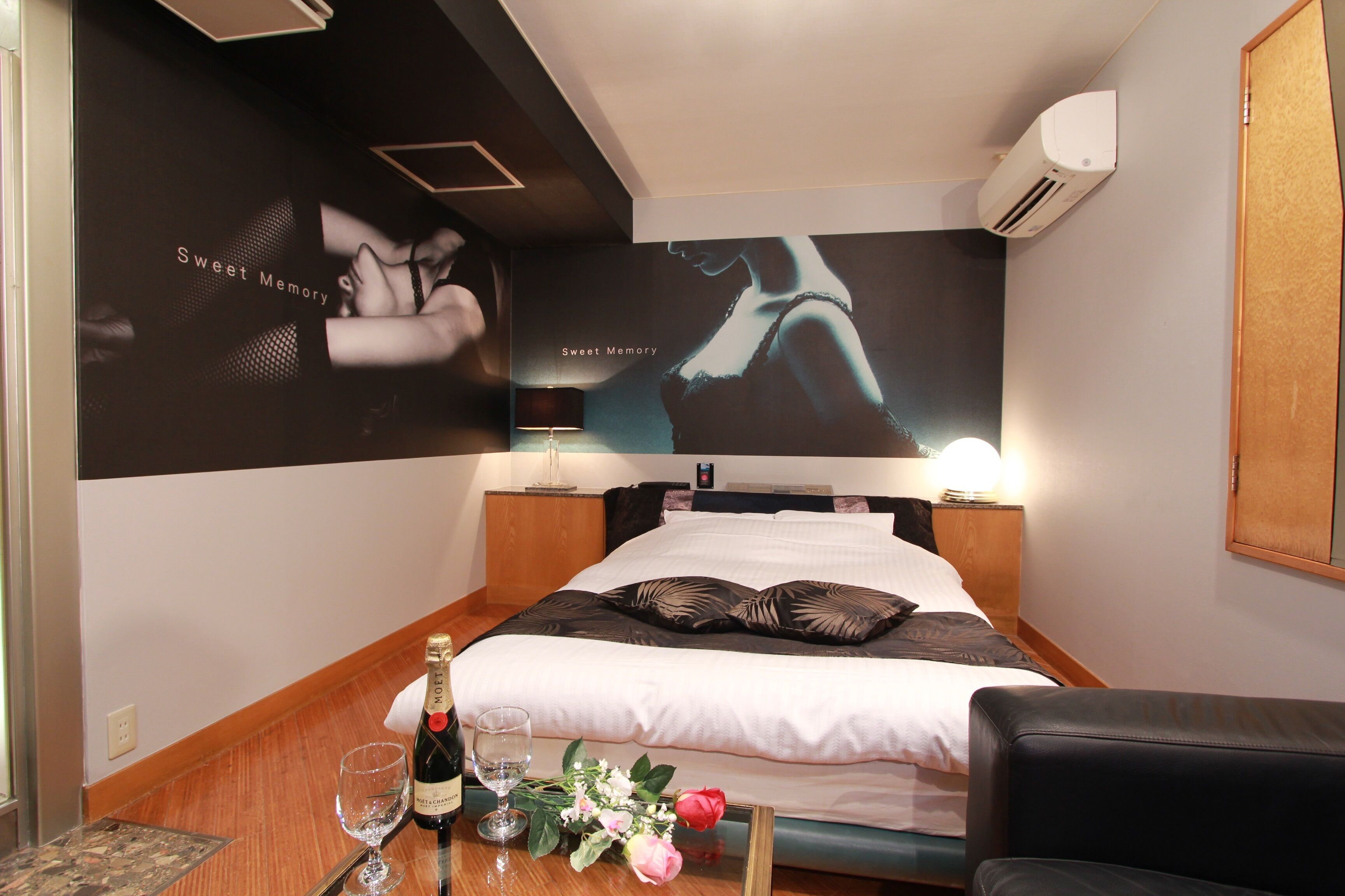Una habitación de hotel con una cama bien hecha, una mesita de noche con una botella de champán, copas y flores, y un cartel grande en la pared.