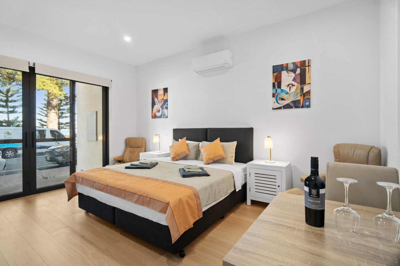 A modern hotel room with a large bed, two bedside tables, a desk with a chair, and a dining table with wine glasses.