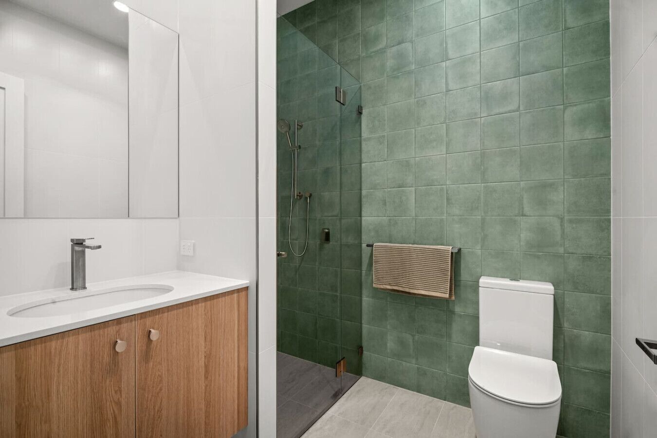 A modern bathroom with a white sink, wooden vanity, green tiled walls, a shower, and a toilet.