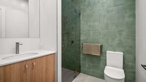 Um banheiro moderno com pia branca, toucador de madeira, paredes com azulejos verdes, chuveiro e vaso sanitário.