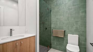 A modern bathroom with a white sink, wooden vanity, green tiled walls, a shower, and a toilet.