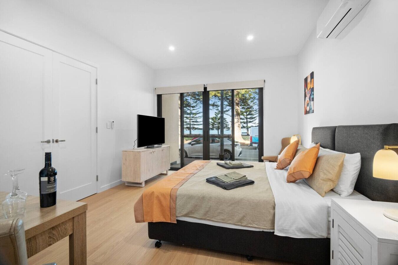 A modern bedroom with a large bed, a TV on a wooden cabinet, a bottle of wine, and a view of the outdoors through large windows.