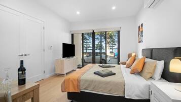 A modern bedroom with a large bed, a TV on a wooden cabinet, a bottle of wine, and a view of the outdoors through large windows.