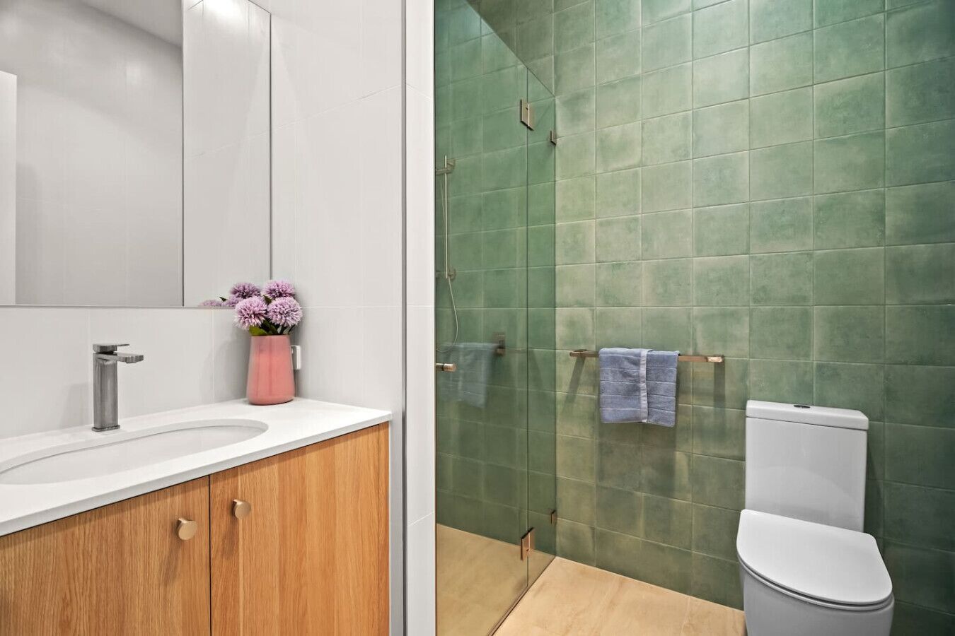 Un baño con un lavamanos blanco, un jarrón rosa con flores y una pared de ducha con azulejos verdes.