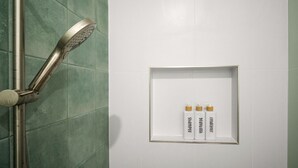 Une douche équipée d’un pommeau de douche à main et d’une commande fixée au mur. On y trouve une étagère blanche sur laquelle sont posées trois bouteilles étiquetées « shampoing », « gel douche » et « après-shampoing ».
