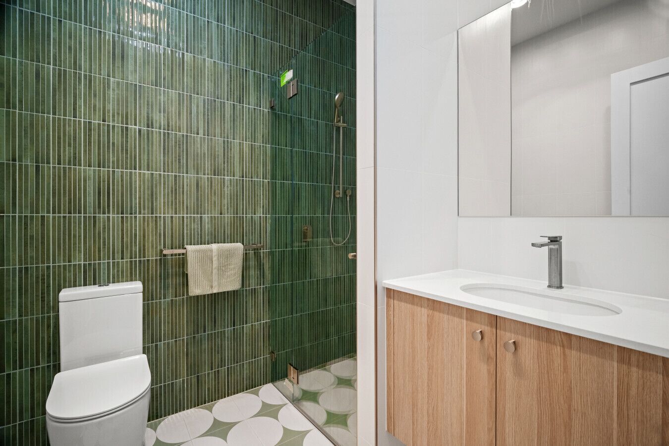 Une salle de bain avec un mur recouvert de carreaux verts, des toilettes blanches, un lavabo avec un meuble en bois, et un miroir au-dessus du lavabo.