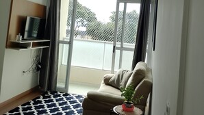 Una sala moderna con un sofá, una mesita con una planta, un televisor en un estante y un amplio ventanal con cortinas.