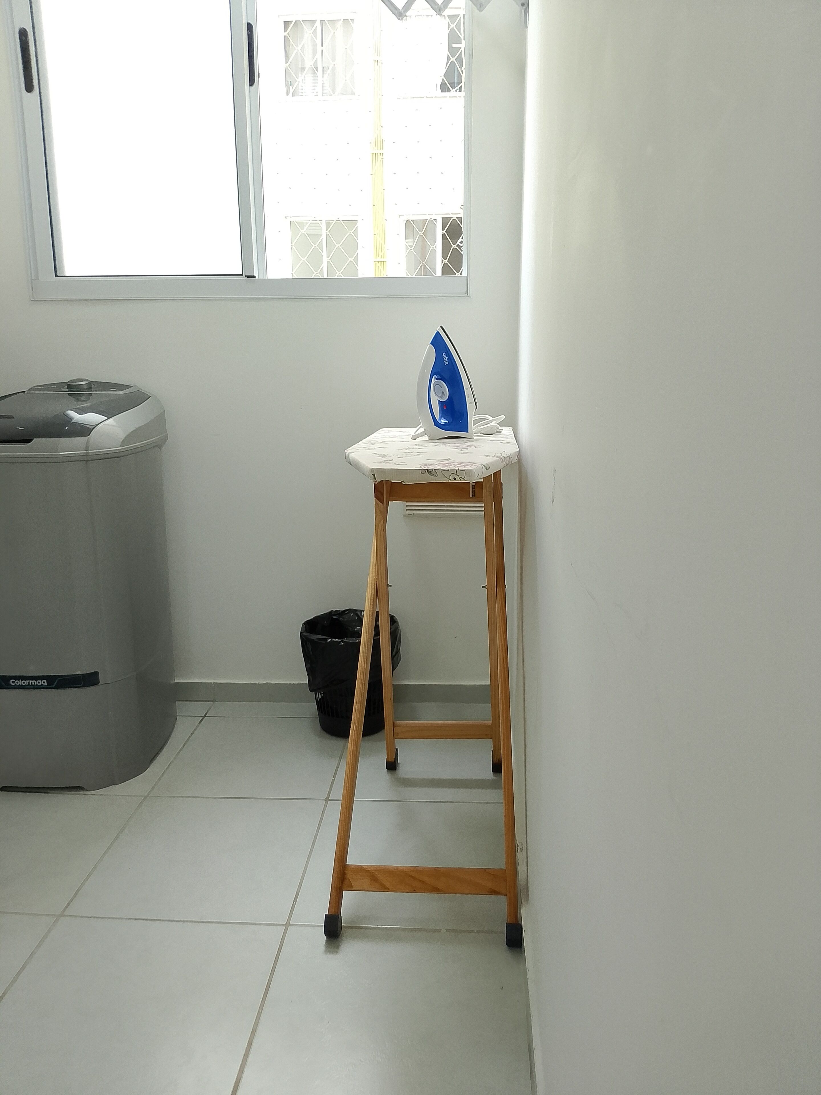 Une petite pièce avec une fenêtre, une poubelle et un tabouret en bois sur lequel est posé un fer à repasser.
