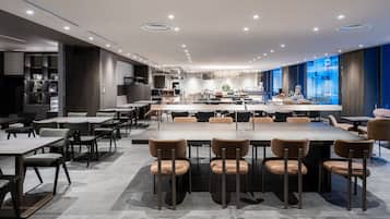 Un moderno comedor con varias mesas y sillas, una cocina al fondo y vista a la ciudad a través de amplios ventanales.