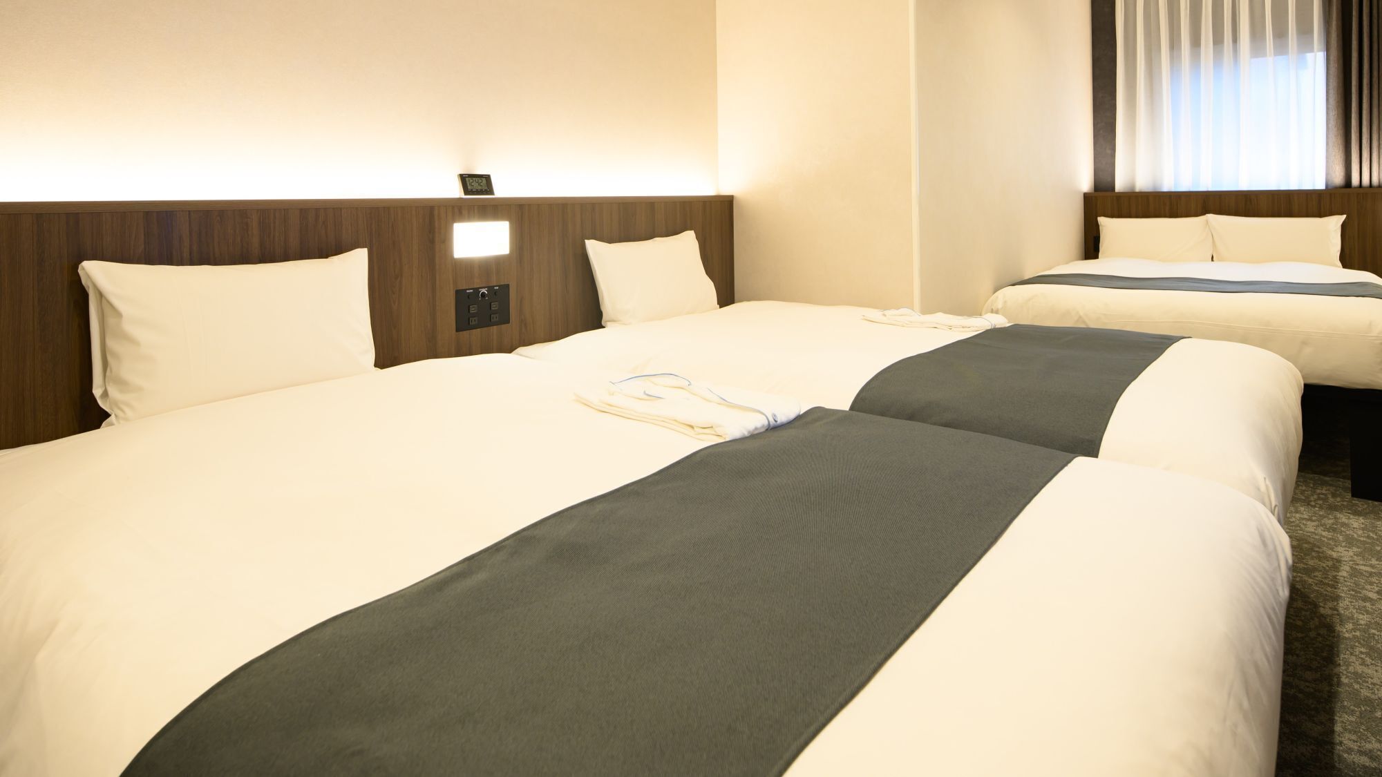 A hotel room with two beds, a nightstand, a wall clock, and a window with curtains.