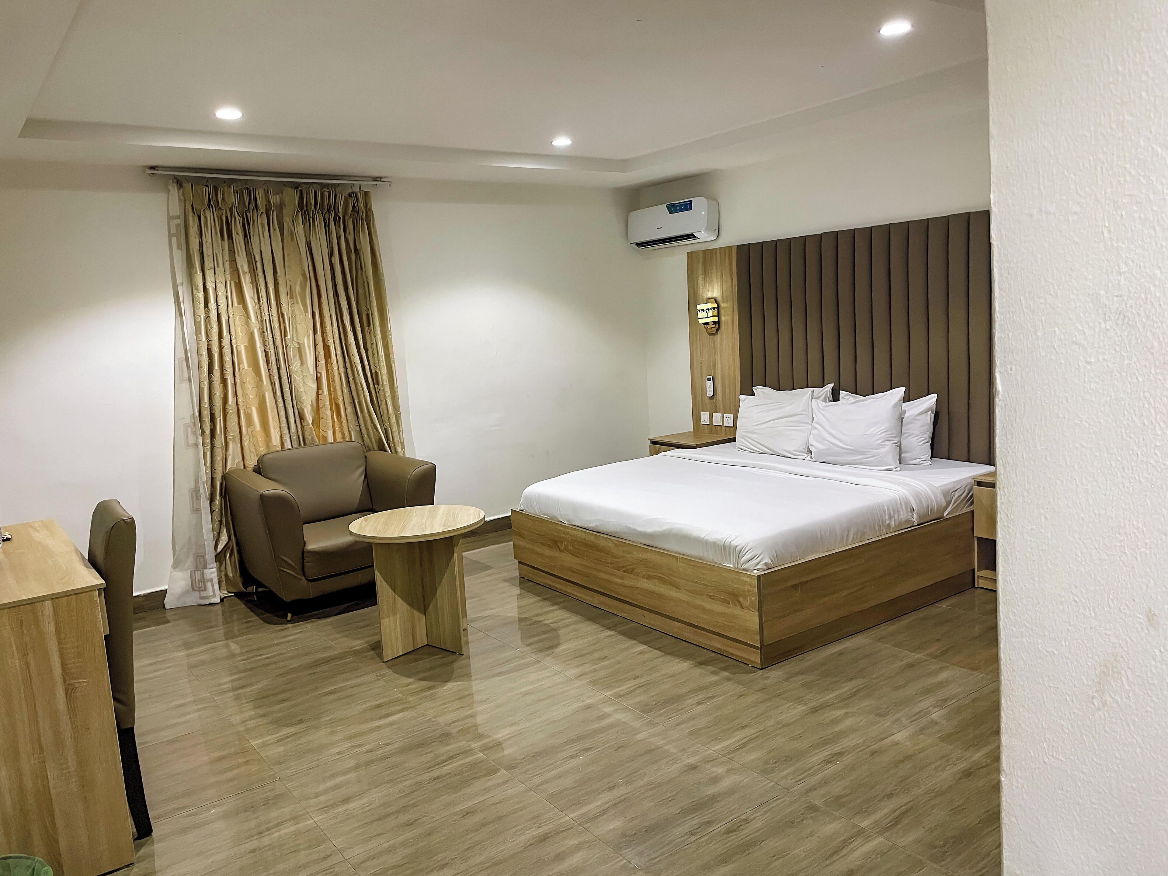 A modern hotel room with a wooden bed, a small round table, a brown leather chair, and a large window with curtains.