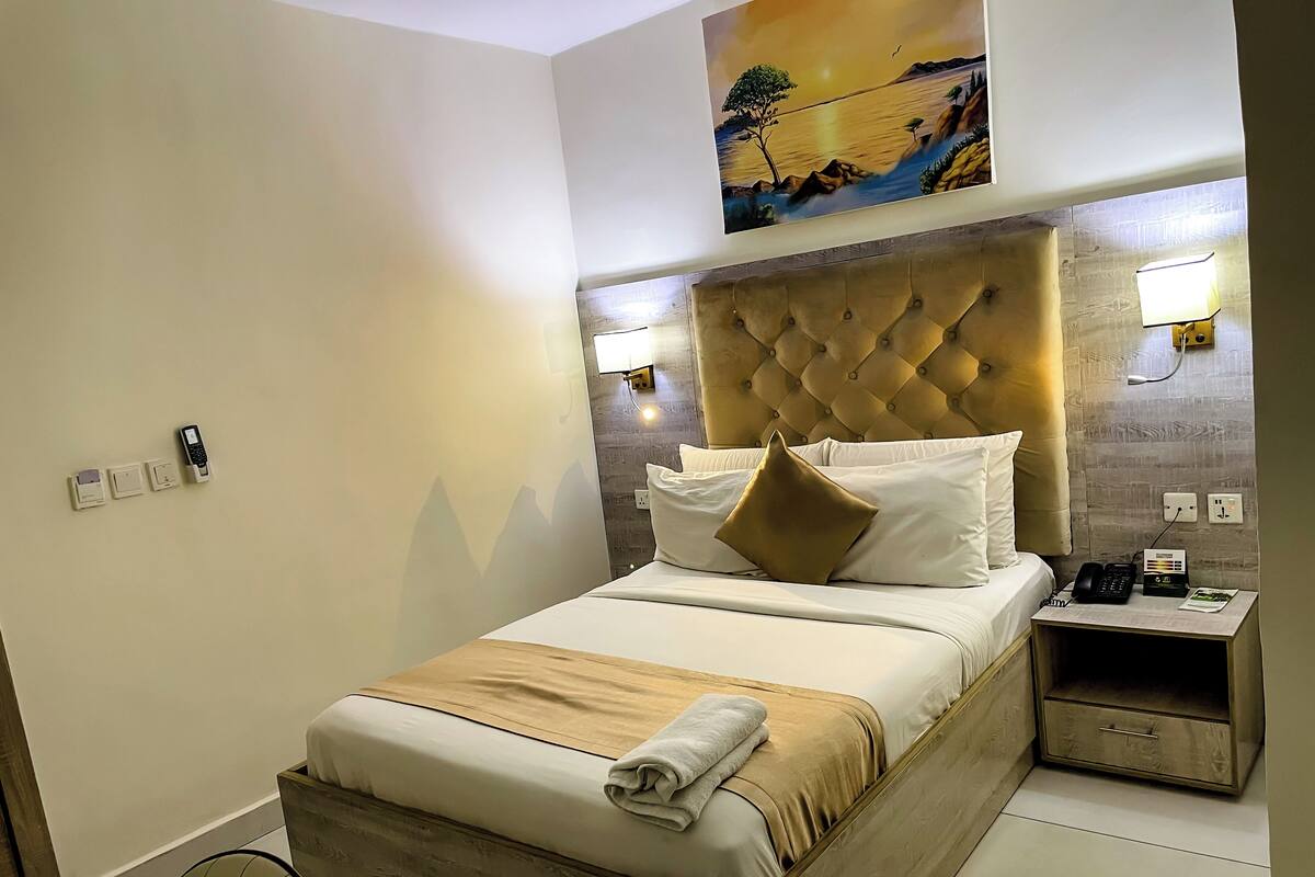 A modern hotel room with a tufted headboard, a bed with white linens and a beige throw, a bedside table with a lamp, and a painting above the bed.