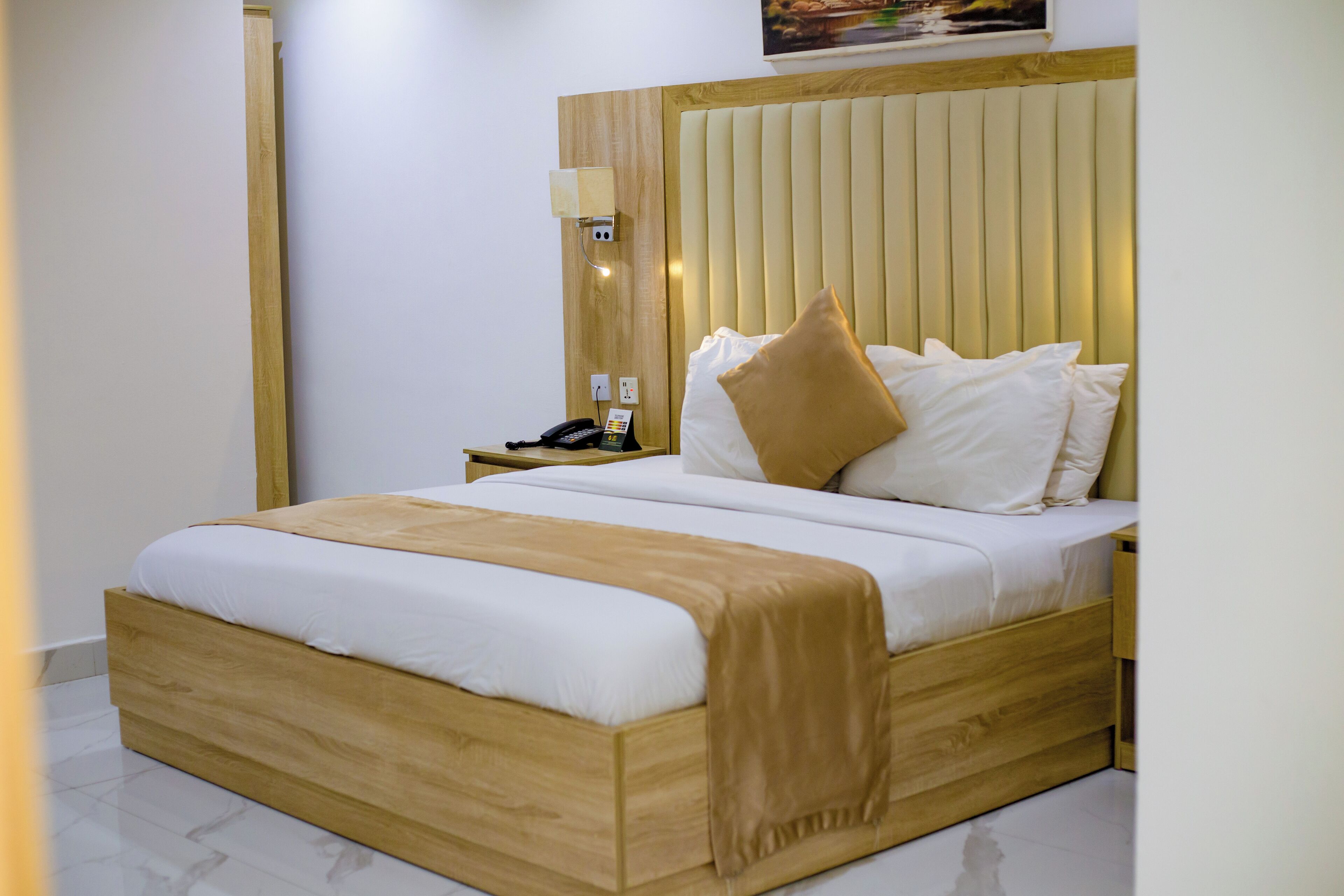 Una habitación de hotel moderna con estructura de cama de madera, ropa de cama blanca, un plaid color beige y una mesita de noche con teléfono y lámpara.
