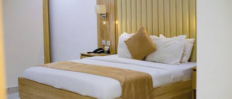 Una habitación de hotel moderna con estructura de cama de madera, ropa de cama blanca, un plaid color beige y una mesita de noche con teléfono y lámpara.