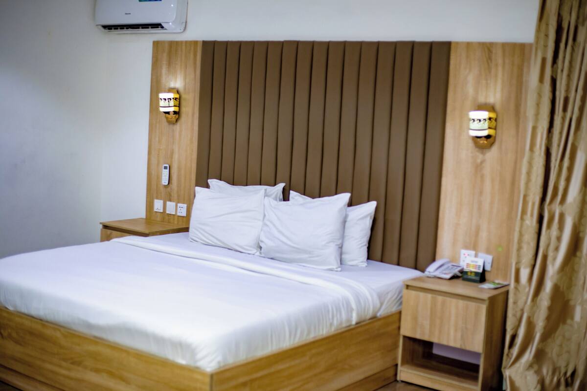 A hotel room with a wooden headboard, white bedding, a bedside table with a phone and a lamp, and a wall-mounted air conditioner.