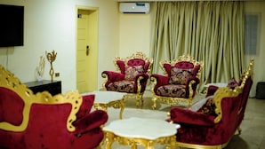 Un salon avec deux fauteuils richement décorés, aux couleurs rouge et or, une table basse blanche et un téléviseur fixé au mur.