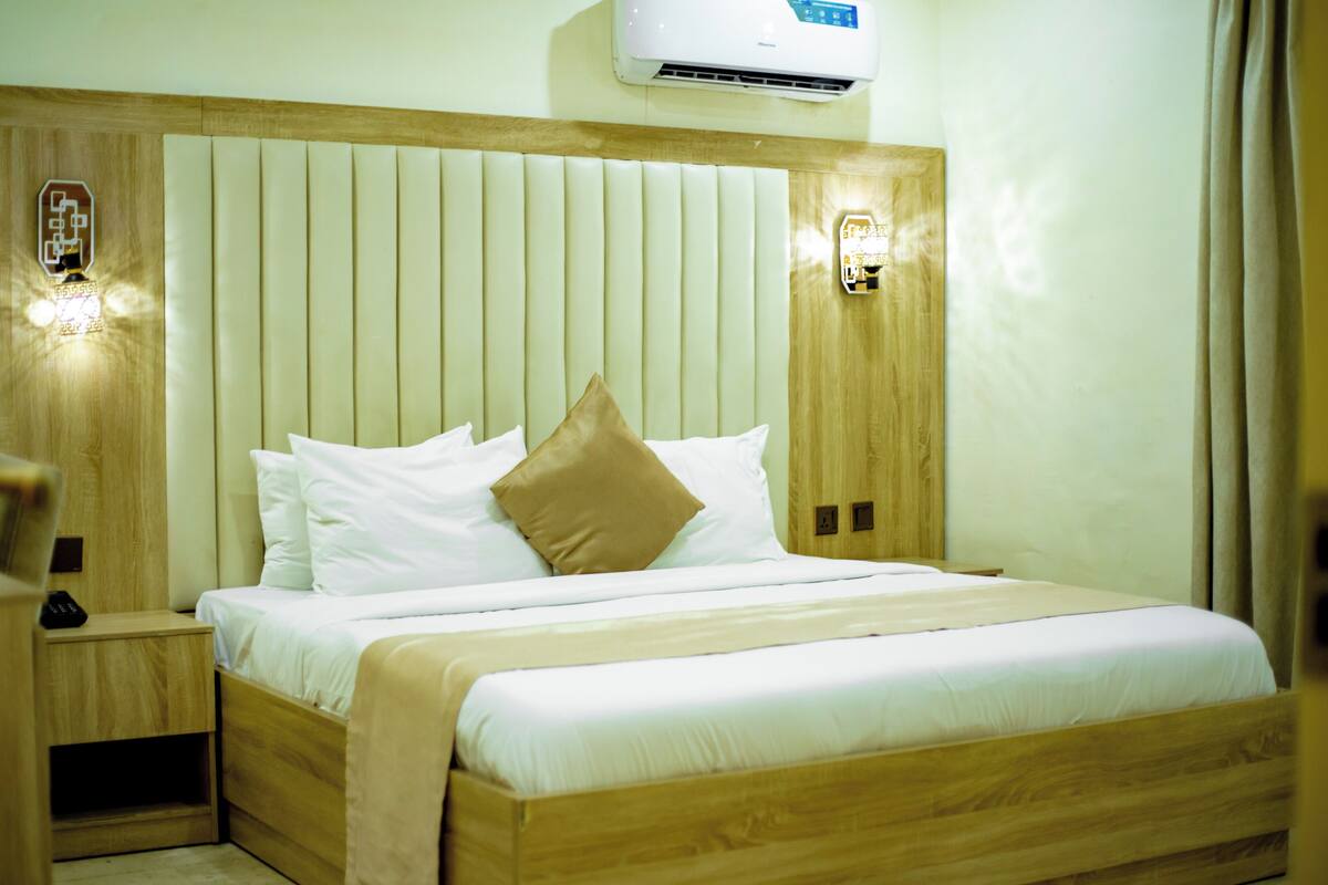 A neatly made bed with white and beige bedding, a wooden headboard, and two wall-mounted light fixtures.