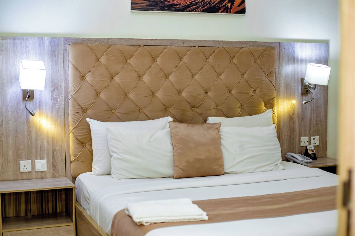 A hotel room with a tufted headboard, white bedding, a brown throw blanket, and bedside lamps.