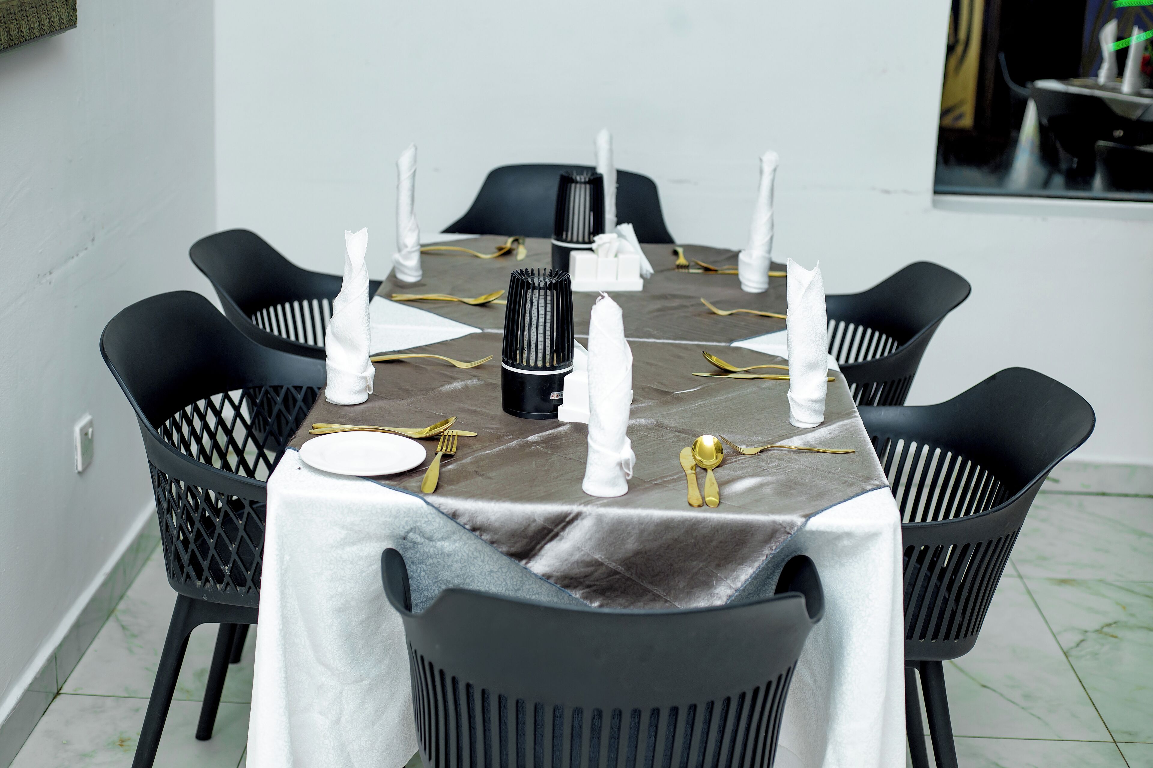 A round dining table with a white tablecloth, black chairs, gold cutlery, and white napkins.