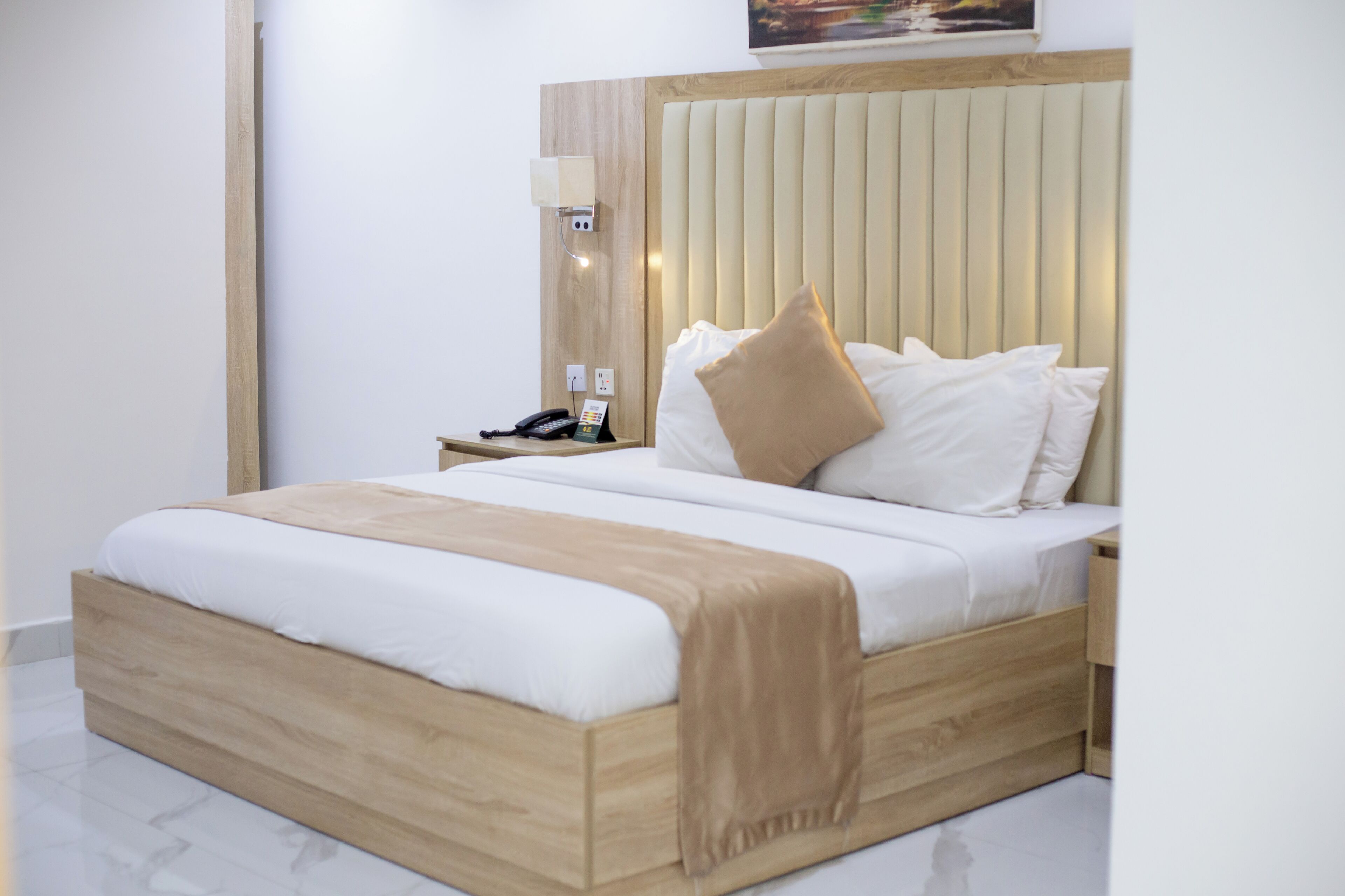 Une chambre d’hôtel moderne avec un cadre de lit en bois, du linge de lit blanc, un plaid beige, une lampe de chevet et une table de chevet avec un téléphone.