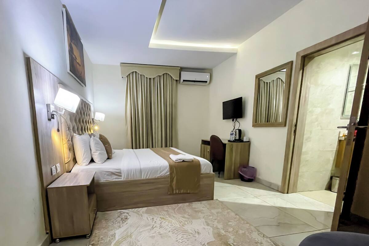 A hotel room with a bed, bedside table, lamp, curtains, TV, and a bathroom door.