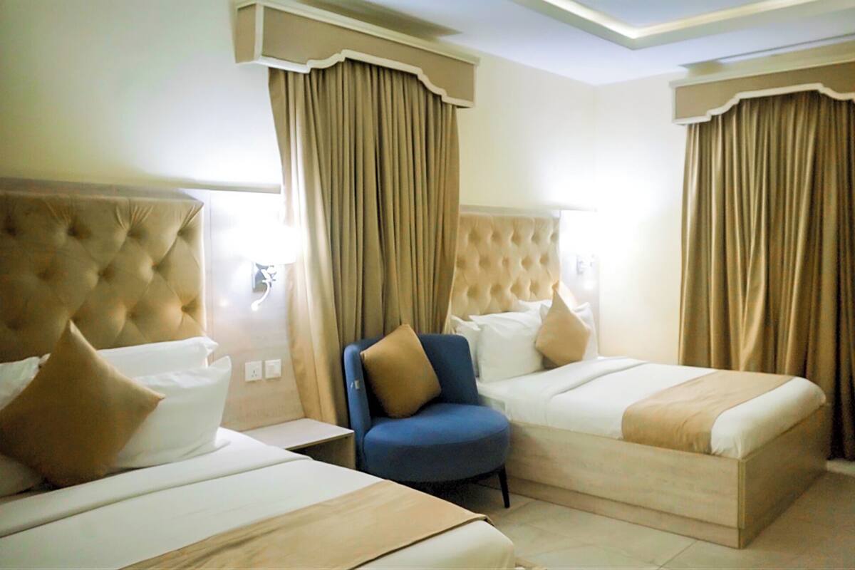 A hotel room with two beds, a blue armchair, and beige curtains.