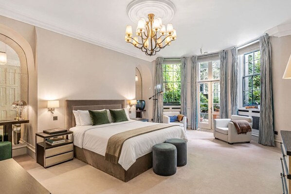 A bedroom with a large bed, bedside tables, a chandelier, a window with curtains, and a view of greenery outside. - Cadogan Square Ultra-luxe 4BR Private Garden (London)