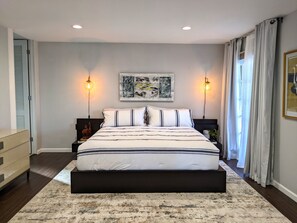 Ein Schlafzimmer mit einem großen Bett, zwei Nachttischlampen, einem gerahmten Kunstwerk über dem Bett und einem Fenster mit Vorhängen.