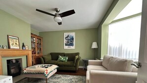 Ein Wohnzimmer mit Kamin, einem grünen Sofa, einem beigen Sessel, einem gemusterten Teppich und einem Deckenventilator.
