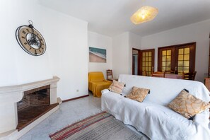 A living room with a fireplace, a sofa with patterned cushions, a yellow armchair, and a wall clock. - Wild Atlantic 2 bedroom apartment (Aguieira)
