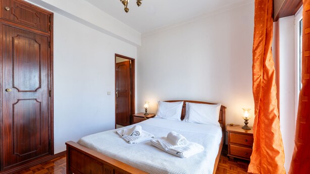 Una cama bien tendida con ropa blanca y toallas, un armario de madera, una mesita de noche con lámpara y suelo de madera.