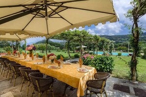 Un área de comedor al aire libre con una mesa larga dispuesta para una comida, rodeada de sillas, bajo un gran paraguas.