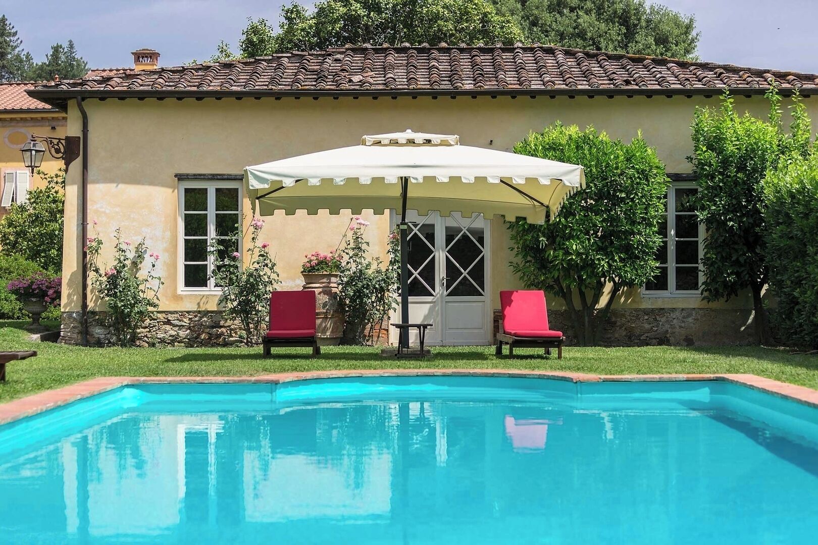 A swimming pool with two red chairs and a white umbrella in front of a yellow building with a tiled roof.