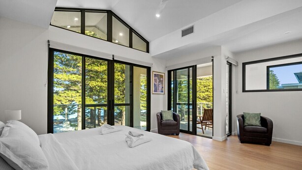 A modern bedroom with a large bed, a balcony with chairs, and a view of trees.