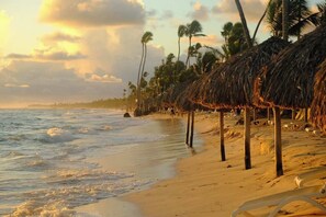 A beach with thatched-roof structures, palm trees, and a sandy shoreline. - New Chic 2BR APT. (Punta Cana)